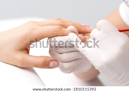 Manicure master in rubber gloves applying transparent nail polish on female nails in beauty salon