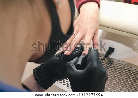 The manicurist in black latex gloves covers the client's nails with a transparent gel polish in the beauty salon.