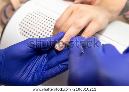 The manicurist cuts the nail cuticles with small scissors, wearing blue latex gloves.