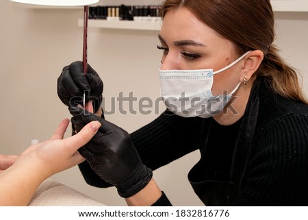 A manicurist in a mask and black gloves paints a client's nails with a brush