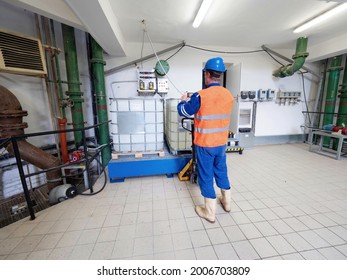 Manipulator In Chemical Operation Wearing Reflective Vest, Blue Helmet And Rubber Work Boots. A Man Handles A Pallet Truck And A Chemical Material Tank.