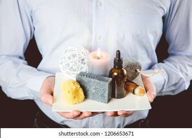 Manly businessman holding white ceramic tray with group of different spa essentials, beard oil, gray soap, natural sea sponge, candle burning, aroma oil on black background. Man spa concept.