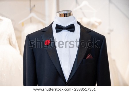 A mannequin dressed in a sharp black tuxedo with a bow tie, white shirt, red rose boutonnière, and pocket square, displayed in a wedding boutique