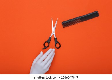 Mannequin Hand Holding Hair Scissors And Comb On Orange Background. Minimalism Beauty Concept. Top View