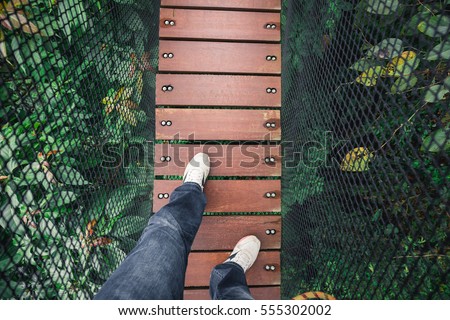 Man's feet and legs on wooden bridge over top of tree for sight seeing. Bridge covered by black net as wall. 