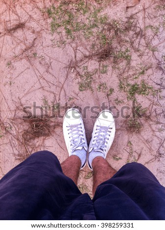 Man's feet are wearing shorts and white shoes standing on the natural ground.
