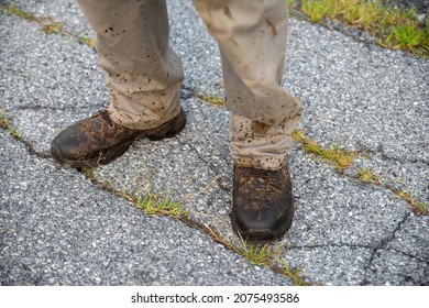 A Man's Feet In Work Boots And Pants Splattered With Dirt And Grease Stands On A Proken Asphalt Road. Natural Light With Copy Space.