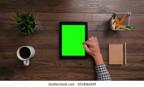 A Man's Finger Scrolling On The Green Touchscreen. The Digital Tablet Is On The Brown Table. View From The Top. Close-up.