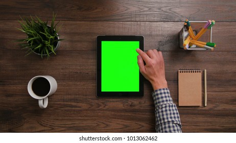 A Man's Finger Scrolling On The Green Touchscreen. The Digital Tablet Is On The Brown Table. View From The Top. Close-up.