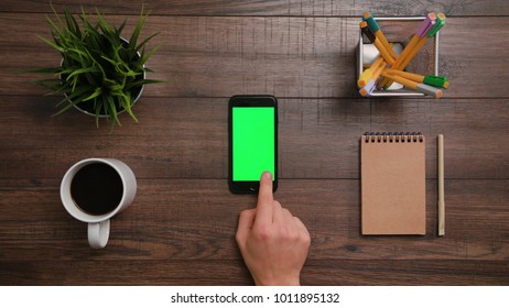 A Man's Finger Scrolling On The Smartphone With The Green Screen. The Phone Is On The Brown Table. View From The Top. Close-up.