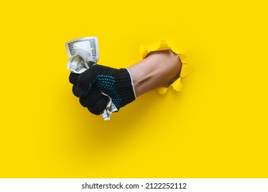 A man's hand in a black fabric work glove holds crumpled dollar bills (money). Torn hole in yellow paper. Concept for gastarbeiter, handyman, salary for job. Copy space.