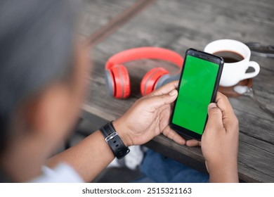 Mans hand in the cafe and using mobile phone green screen. There's a red head phone and coffee on his desk.. Lifestyle, business, technology concept. copy space for advertisement writing