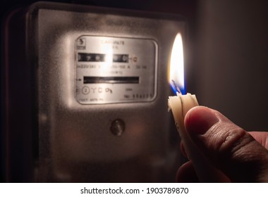 A man's hand with candle in complete darkness near an electricity meter at home. Power outage, blackout, non-payment of electricity bill concept.