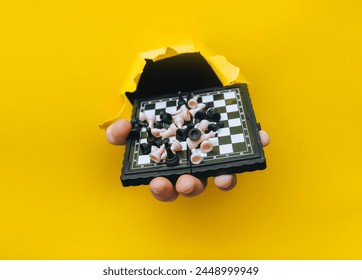 A man's hand comes out of a torn hole in yellow paper and holds a small chessboard with pieces scattered on it. The concept of intellectual development and games that develop thinking. - Powered by Shutterstock