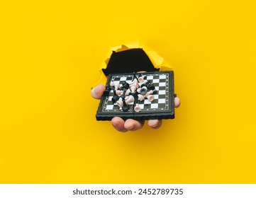 A man's hand comes out of a torn hole in yellow paper and holds a small chessboard with pieces scattered on it. The concept of intellectual development and games that develop thinking. - Powered by Shutterstock