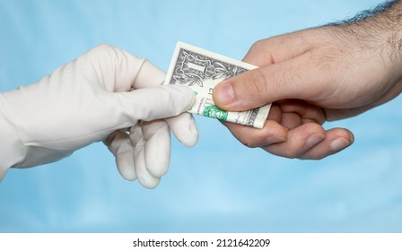 man's hand is giving a dolar to a hand in white surgical glove, nurse or doctor. the hand in glove takes the money. corruption in medicine field. pandemic times. blue medicinal background.