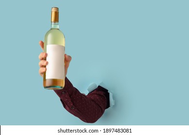 Man's Hand Holding Bottle Of Wine And Breaking Through Blue Paper Background.