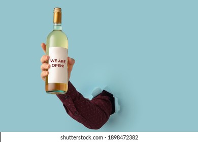 Man's Hand Holding Bottle Of Wine And Torn Through Blue Paper Background.