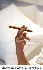 Mans hand is holding a cigar. The focus is on the hand and the cigar, with the background blurred. The fingers delicately position around the cigar, highlighting its texture and form. Smoking cigar