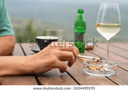A man's hand is holding a cigarette between his two fingers. The background is blurry.