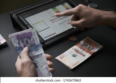 A Man's Hand Holding Colombian Bills While He Does The Math On A Tablet