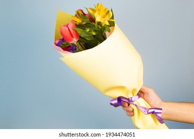 Man's Hand Holding A Decorated Bouquet Of Flowers. Man Giving Flowers In Yellow Wrapping Paper, Indoors.