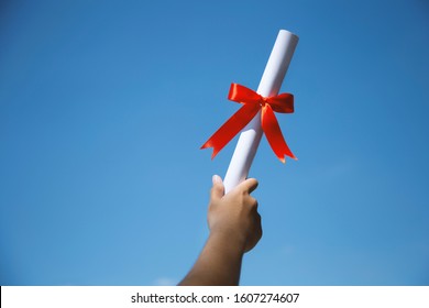 Man's Hand Holding A Diploma With A Red Ribbon On Blue Sky Background