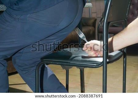 A man's hand holding a fork over a chair that another person sits on. Joke or malicious intent. An example of hemorrhoid pain.                              