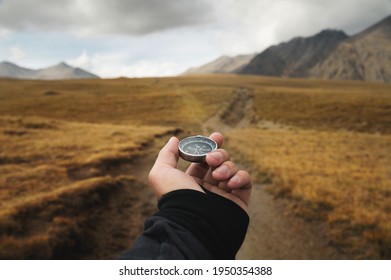 Man's hand holding a magnetic compass first-person view against the background of a high-altitude path and mountains - Shutterstock ID 1950354388