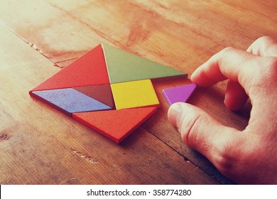 man's hand holding a missing piece in a square tangram puzzle, over wooden table. 