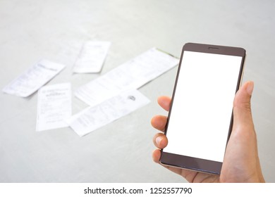 Man's Hand Holding Mobile Phone With White Screen And Utility Bills Or Slips On Gray Table Blurred Background