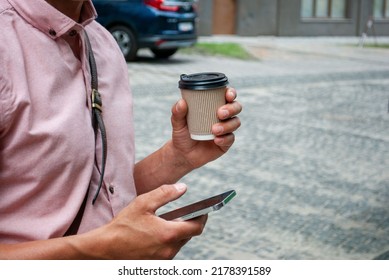 Man's Hand Holding Mobile Phone With A Paper Craft Cup Of Coffee On Street. Close Up Of Man Hands With Cup And Telephone In Hands