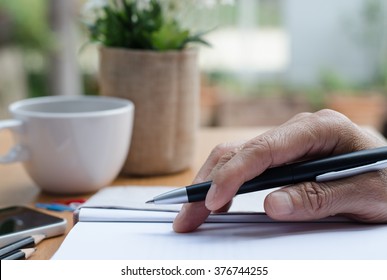 Man's Hand Holding A Pen On Paper Notebook On Table In Relax Position.