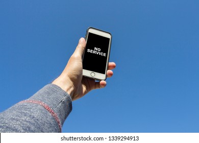 Man's Hand Is Holding A Phone And Trying To Find A Connection Against The Blue Sky. On The Phone Screen, The Symbol With No Signal