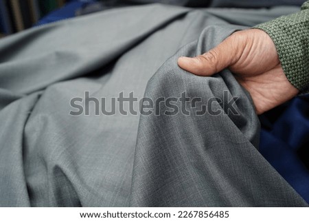 A man's hand holding a piece of quality silver grey fabric. Choosing a textile material in a tactile and visual way to tailor a garment, trouser, jacket. Close-up, selective focus, blurred background.