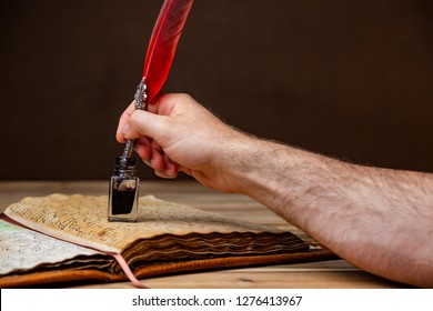 A Mans Hand Holding A Red And Silver Quill Pen And Dipping Ink On An Old Book Resting On A Wooden Table.