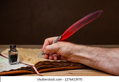 A Mans Hand Holding A Red And Silver Quill Pen And Writing In An Old Book Resting On A Wooden Table.