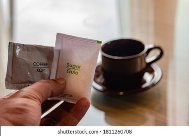 Man's Hand Is Holding A Sachet Of Sugar And Instant Coffee With A Blurred Background Of A Dark Coffee Cup.