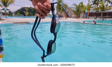 Man's Hand Holding Swimming Goggles With A Swimming Pool In The Background, The Concept Of Sports.
