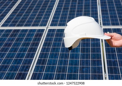 Man's Hand Holding White Protective Cask, Hand Close, Solar Pannel Field Background. Hot Sunny Summer Weather. Copy Space. Concept Of Renewable And Clean Energy, Technology.