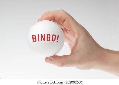 Man's Hand Holding White Styrofoam Ball Against The White Background With Bingo Written Inside It. 