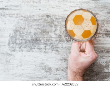 Man's Hand Holds A Beer Mug With A Football On A Beer Foam. Top View. Space For Text