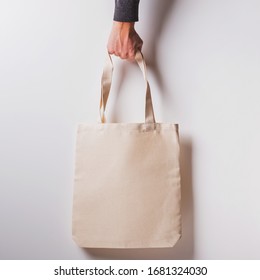 Man's Hand Holds Blank Cotton Eco Tote Bag Over The White Wall, Design Mockup.