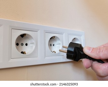 A man's hand holds an electrical plug next to the sockets on the wall.