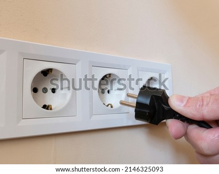 A man's hand holds an electrical plug next to the sockets on the wall.