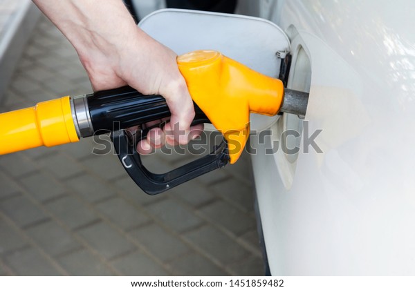 A man\'s hand holds a filling gun inserted into\
the hole of a gasoline tank of a car on a gasoline fueling.\
Close-up of the hand and fuel filling pistol. Yellow refueling gun\
at the refuel station.