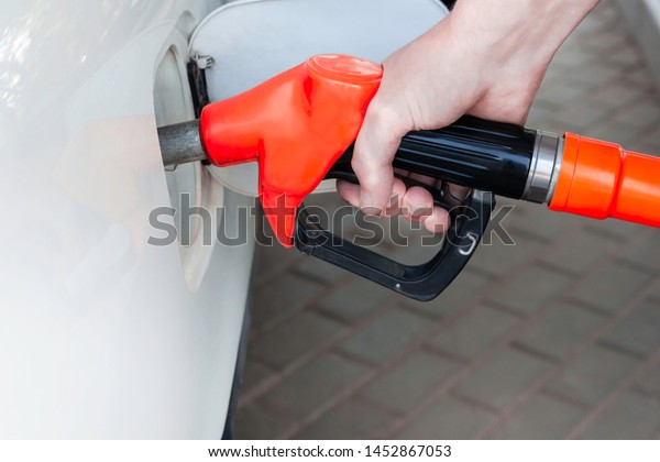 A man\'s hand holds a filling gun inserted into\
the hole of a gasoline tank of a car on a gasoline fueling.\
Close-up of the hand and fuel filling pistol. Red refueling gun at\
the refuel station.