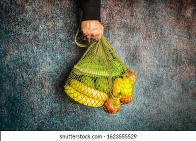 A Man's Hand Holds A Green String Bag With Fresh Vegetables And Fruit On A Dark Background. No Plastic, Only Natural Materials And Natural Products.