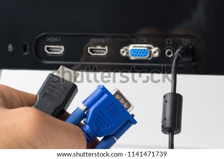 Man`s hand holds HDMI and VGA cables against a monitor with ports. Choise between modern HDMI and old VGA connection