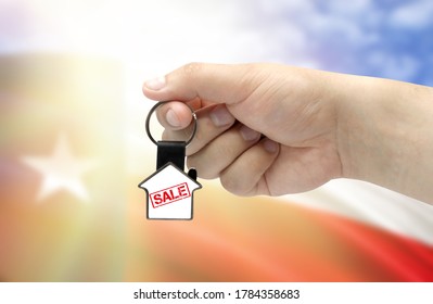 A Man's Hand Holds A Keychain In The Form Of A House With The Inscription Sale On The Background Of The Flag Of State Of Texas. Property And Real Estate Sale Concept.